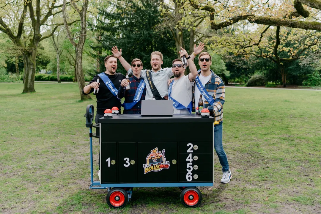 Fünf Männer mit Team-Groom-Schärpen jubeln hinter dem Battle-Wagen im Park
